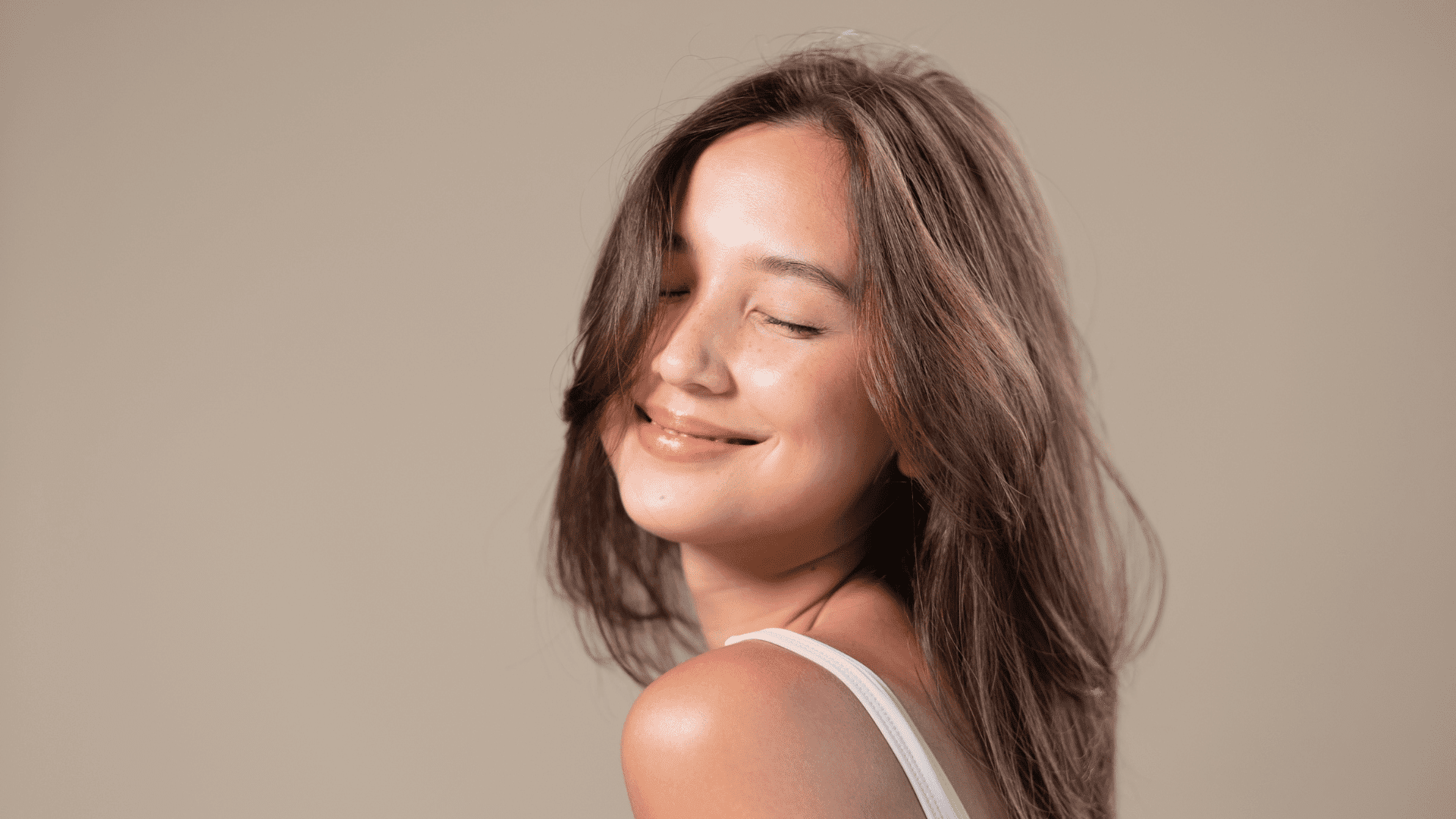 Smiling woman with closed eyes, long brown hair, in a white top against a plain background.